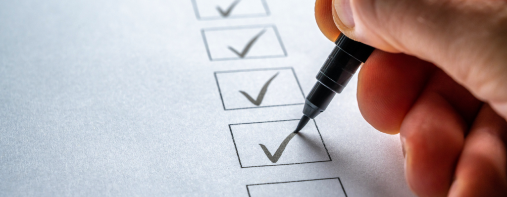 Mão marca itens em uma lista de verificação, representando organização, acompanhamento de tarefas e a importância de delegar responsabilidades na gestão de equipes.
